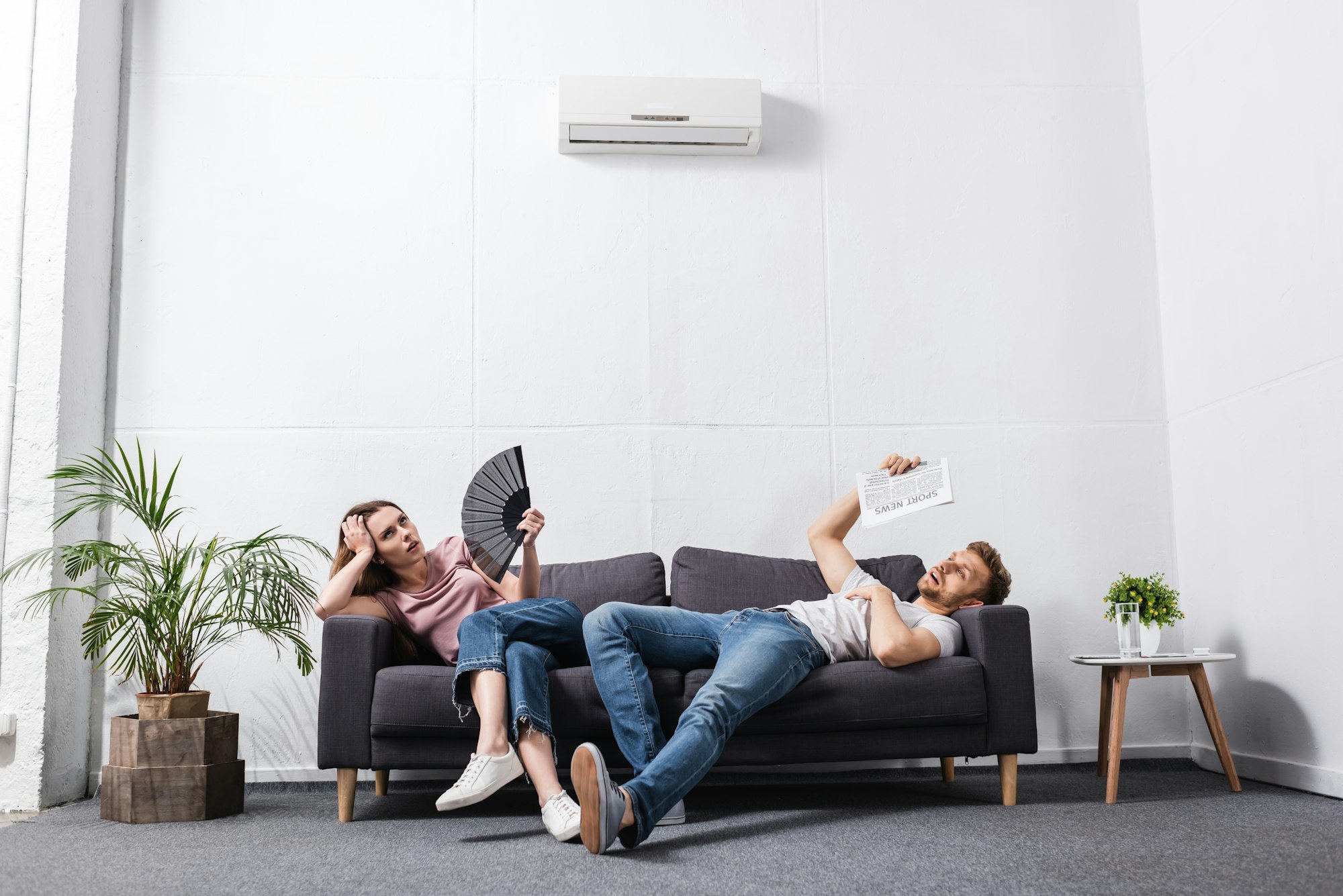 young couple with hand fan and newspaper suffering from heat at home with broken air conditioner